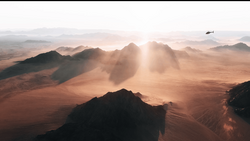 Aerial view of a desert landscape with mountains silhouetted against golden sunlight and dust clouds.
