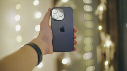 Hand holding a dark blue iPhone with quad camera setup against blurred warm-toned bokeh background.