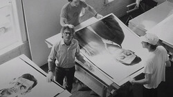 Black and white overhead view of four people examining large photographic prints on a work table.