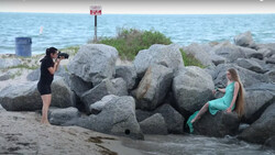 Photographer shooting a mermaid-tailed model posed on large boulders at a beach.