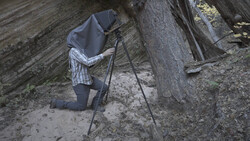 Photographer kneeling behind tripod-mounted camera in rocky cave setting, with dark cloth covering head and camera.