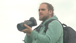 Male photographer holding a DSLR camera with telephoto lens, wearing a green jacket and backpack.