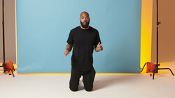 Man in black clothing kneeling on white floor against colorful backdrop with geometric shapes.