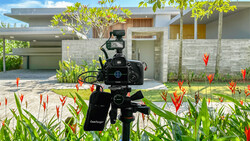 Professional video camera on tripod filming a modern residential house surrounded by tropical plants.
