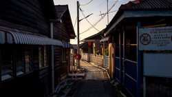 Narrow alley lined with dark wooden buildings at dusk, with warm golden light illuminating the street and distant figures walking.