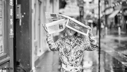 Person wearing patterned shirt holds newspapers above their head on a city street.