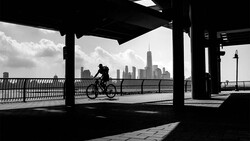 Silhouetted cyclist riding on a waterfront path beneath a bridge structure, with a city skyline in the background.