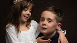 Young girl in white dress with tiara and boy in dark shirt posed together against dark background.