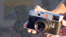Hands holding a silver rangefinder camera with a dark telephoto lens in the background, backlit by warm golden light.