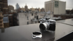 Vintage rangefinder camera with lens cap displayed on a rooftop overlooking an urban cityscape.