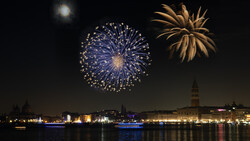 Blue and gold fireworks exploding over a waterfront cityscape at night with illuminated buildings reflected in calm water.