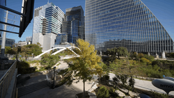 Modern urban plaza with curved white architectural feature, high-rise office buildings, and mature trees under clear blue sky.
