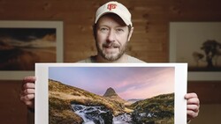 Photographer displaying a landscape print of a mountain peak with stream and valley at golden hour.