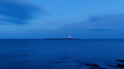 Lighthouse beacon glowing on a distant island at twilight over calm blue ocean.