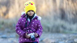 Child in purple jacket and yellow Pikachu beanie holding a camera outdoors.