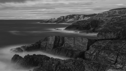 Rugged coastal cliffs meet misty ocean waters under stormy skies in black and white.