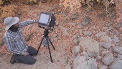 Cinematographer operating a monitor mounted on a tripod while filming in a rocky desert landscape.