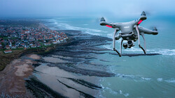Drone with camera flying above coastal cliffs, sand patterns, and seaside town with ocean in background.