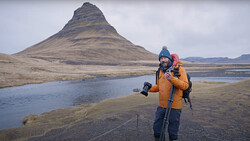 Photographer in yellow jacket with camera and tripod standing in Icelandic landscape with cone-shaped mountain across water.