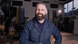 Bearded man in denim jacket smiling in a professional studio setting with lighting equipment visible.