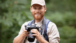 Smiling photographer in white cap holding DSLR camera outdoors with blurred green foliage background.