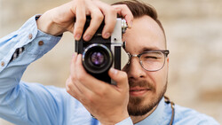 Photographer wearing glasses holding vintage film camera to his eye while looking through viewfinder.