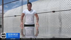 Man in white t-shirt and light pants standing against chain-link fence with blue sky background.