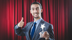 Professional man in business suit holding star-shaped award with thumbs up gesture against red curtain backdrop.