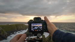 Photographer holding a mirrorless camera displaying a coastal landscape on its rear screen during golden hour.