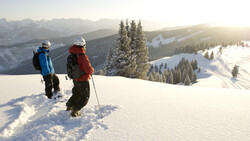 Two hikers in winter gear walking through deep snow on a mountain slope with evergreen trees and snow-covered valleys visible below.