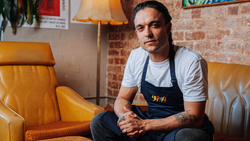 Man in dark apron seated on mustard-yellow leather chair against exposed brick wall.