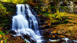 Cascading waterfall flows over rocky ledges surrounded by autumn foliage and moss-covered stone.