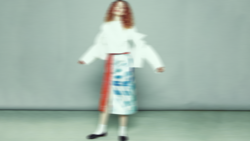 Woman in white cardigan and blue patterned skirt poses with arms extended against neutral background.