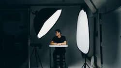 Photographer seated at desk in studio with two large oval softbox lights positioned on either side.
