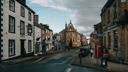 Scottish street lined with historic stone buildings and a red telephone box, sloping downward toward a church spire in the distance.