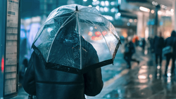 Person holding a clear umbrella during rainfall in an urban street at night with blurred city lights in background.