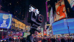 Action camera mounted on tripod in crowded urban plaza at night with illuminated billboards surrounding it.