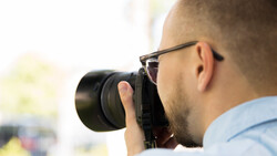 Photographer wearing glasses holding a DSLR camera to his face while shooting outdoors.