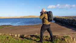 Photographer with camera and backpack standing on a beach, photographing the coastline and hills.
