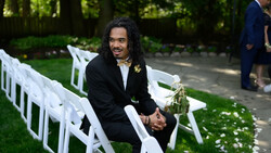 Groom in black suit smiling while seated on white chair at outdoor wedding ceremony.