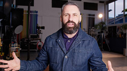 Man with beard in denim jacket gesturing in a professional studio with lighting equipment visible in background.