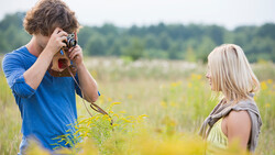 Young photographer holding vintage camera while photographing blonde woman in wildflower field.