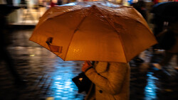 Person holding a golden-orange umbrella on a wet street at dusk, with blurred pedestrians and reflective pavement in the background.
