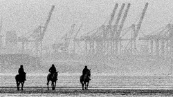 Three silhouetted figures on horseback facing an industrial waterfront with cranes and shipping infrastructure shrouded in mist.