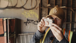 Photographer holding a vintage film camera while wearing a baseball cap and rainbow-striped lanyard in an industrial setting.