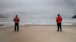 Two people in red jackets standing separately on a beach facing the ocean under overcast skies.
