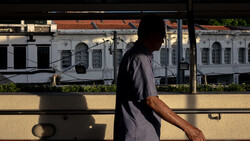 Silhouetted figure leaning against a railing with colonial-style buildings visible through glass in bright sunlight.