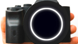 Compact camera with black body and orange grip held in hand, featuring illuminated circular lens ring.