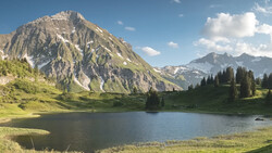 Alpine lake surrounded by coniferous forest with dramatic rocky mountain peak and green meadow.