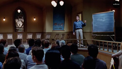 Man presenting to seated audience in wood-paneled room with blue whiteboard behind him.